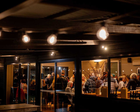 People gathered inside Bangor Vineyard Shed at night with large windows, and festoon lighting.