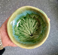 A handmade ceramic bowl with a glossy finish, featuring a leaf design in shades of green and blue on a yellow-beige base, held in a hand against a gray textured background.
