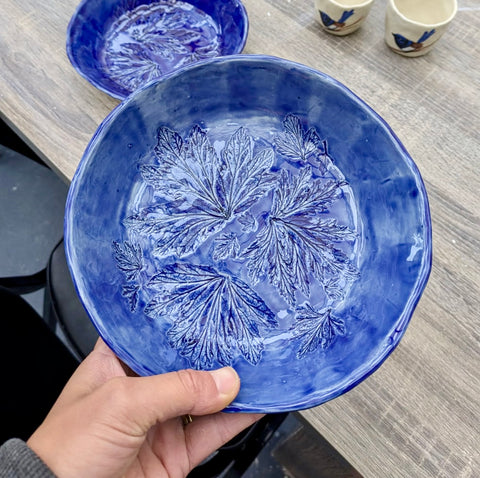 A hand holds a blue ceramic bowl with embossed floral patterns, with other ceramic dishes visible in the background on a wooden table.
