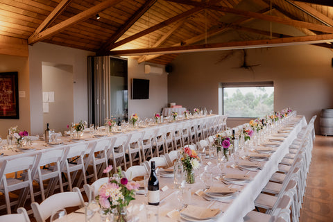 Long table set for a wedding with chairs, flowers, and wine bottles in a room with wooden beams. Bangor Vineyard Tasmania.